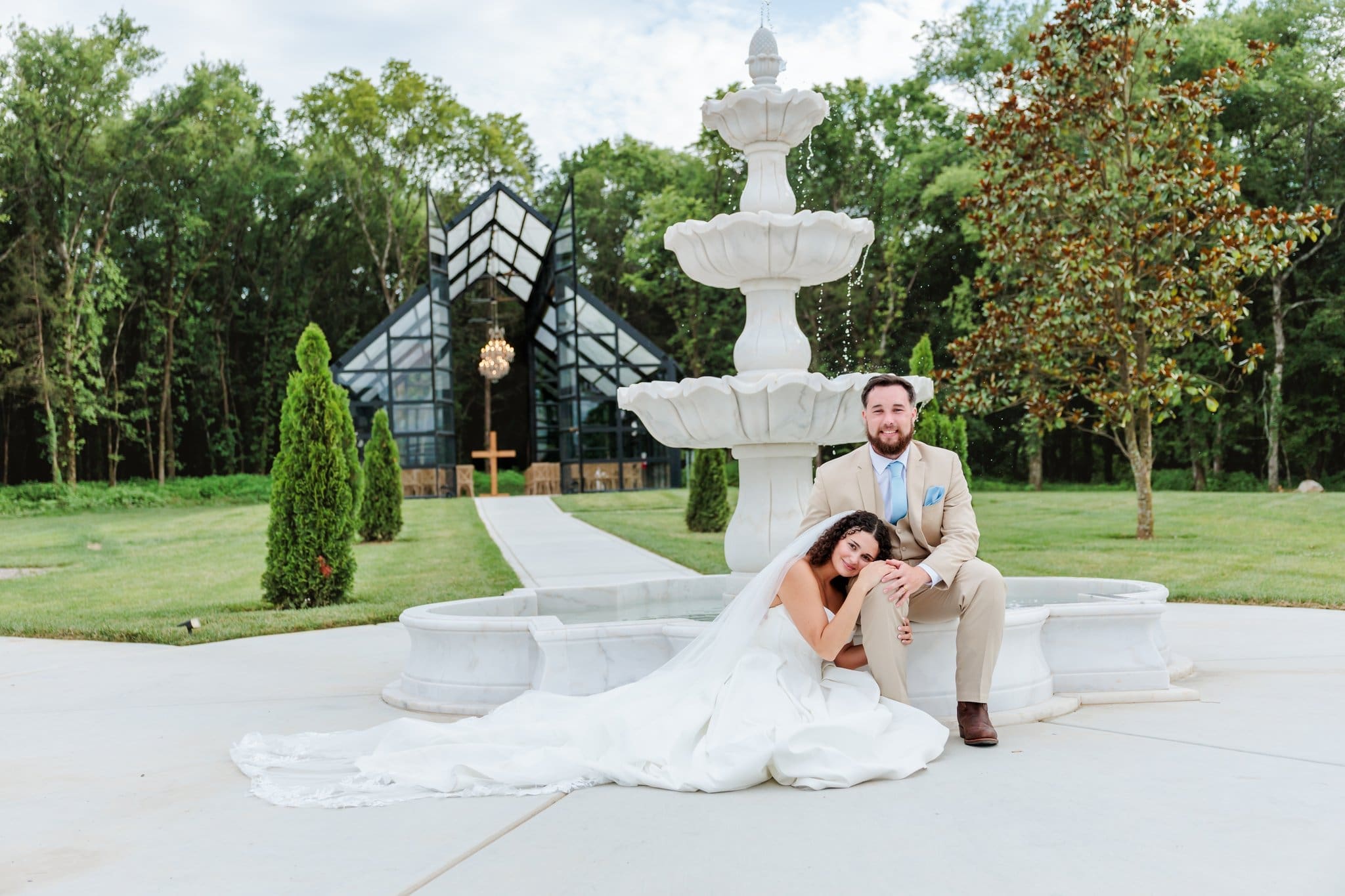 Bride laying her head on her Groom's lap in front of a glass chapel and white marble fountain.
